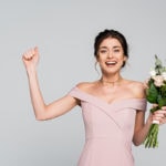 Excited bridesmaid in pale rose dress showing win gesture and holding a bouquet.
