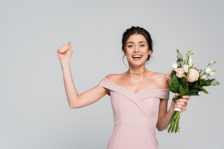 Excited bridesmaid in pale rose dress showing win gesture and holding a bouquet.